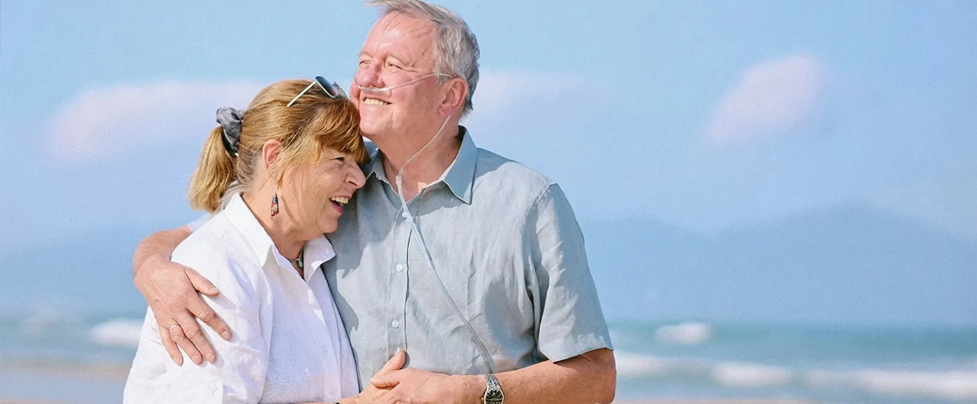 Senior couple hugging and smiling on a beach with a clear blue sky. The man is using a nasal cannula.