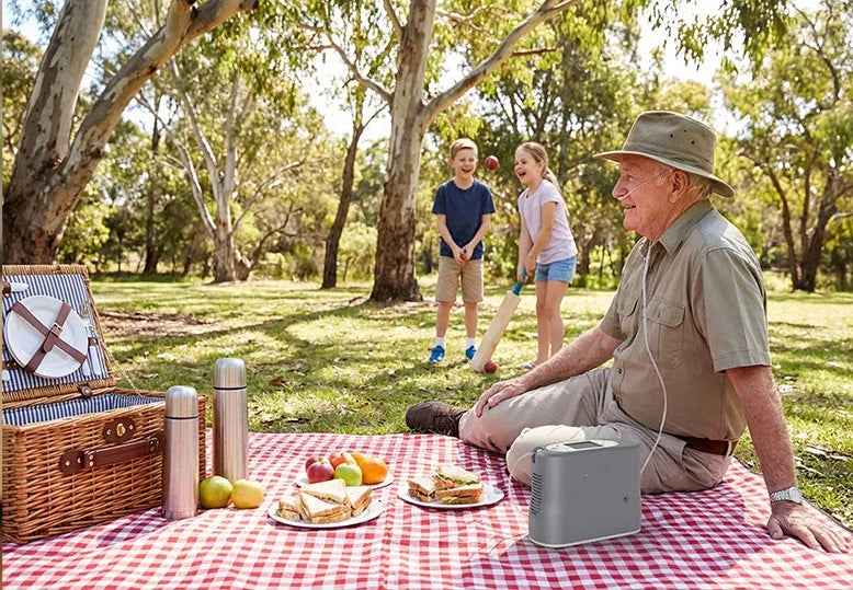 Old man using a nasal cannula on a picnic with his grandchildren in the background playing cricket.