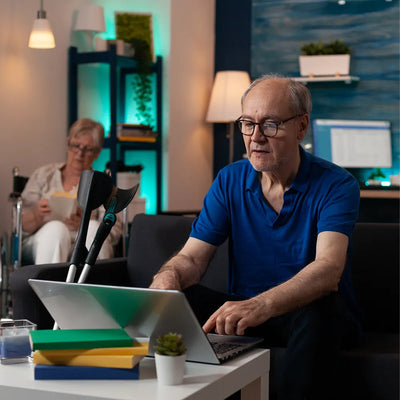 Old man using a laptop in a home office setting with another person in the background.